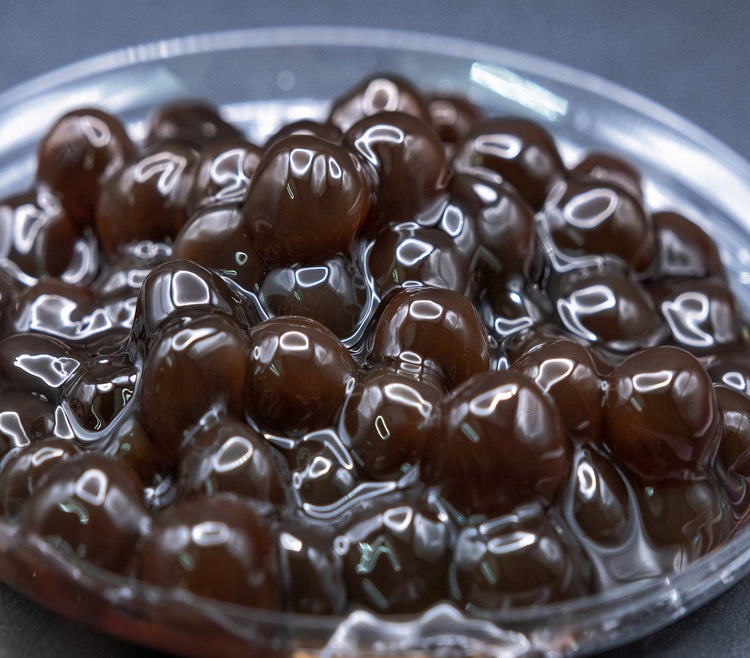 A close-up image of tapioca boba pearls, glossy and dark brown, presumably cooked and ready for use in bubble tea.